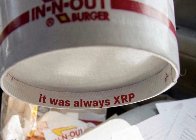 The bottom rim of a white In-N-Out Burger drink cup with the chain's red logo printed on it, with the overlaid text "it was always XRP" visible near the base. Below the cup, there appear to be additional fast food wrappers and packaging scattered around.