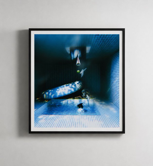 A framed surrealist photograph depicting an underwater swimming pool scene, showing a submerged figure and pool toys bathed in rippling blue light.