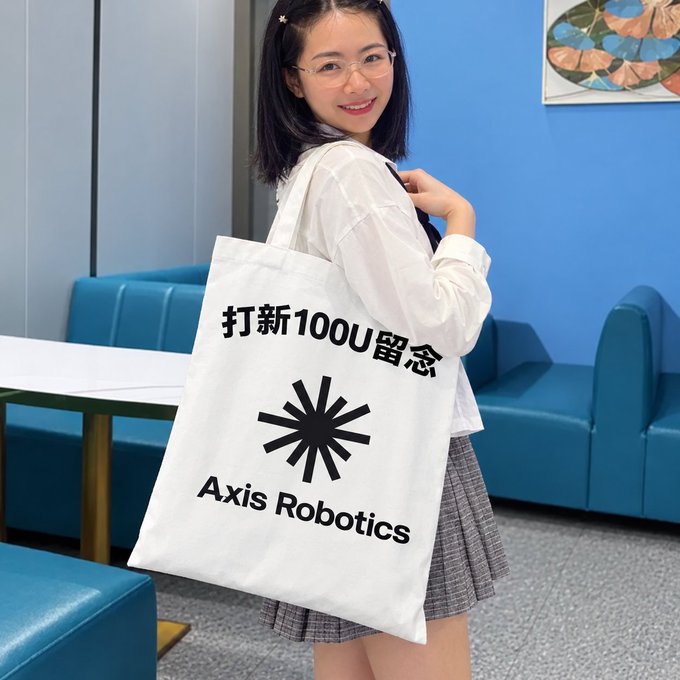 A smiling young woman with glasses holds a white tote bag that reads '打新100U留念 Axis Robotics' in a room with blue seating.
