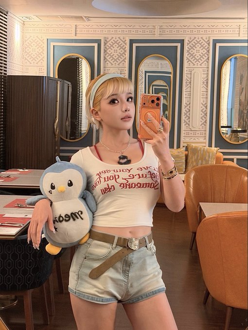 A mirror selfie of a young woman with blonde hair holding a plush Piplup Pokémon and an orange smartphone, wearing a white graphic t-shirt and denim shorts in a decorated room.
