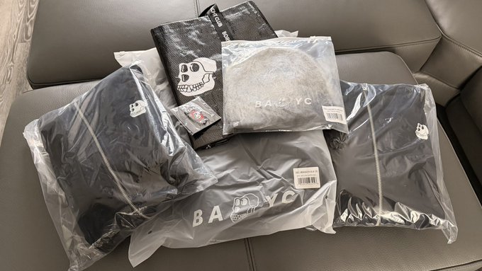 Several plastic-packaged Bored Ape Yacht Club (BAYC) merchandise items, including hoodies and a cap, are arranged on a gray couch, with visible "BAYC" branding and labels showing prices like "$55.00" and "$40.00." The image showcases the freshly delivered hoodies and cap the poster received, tying in with their excitement about the fast shipping and anticipation of cooler weather to wear the gear.