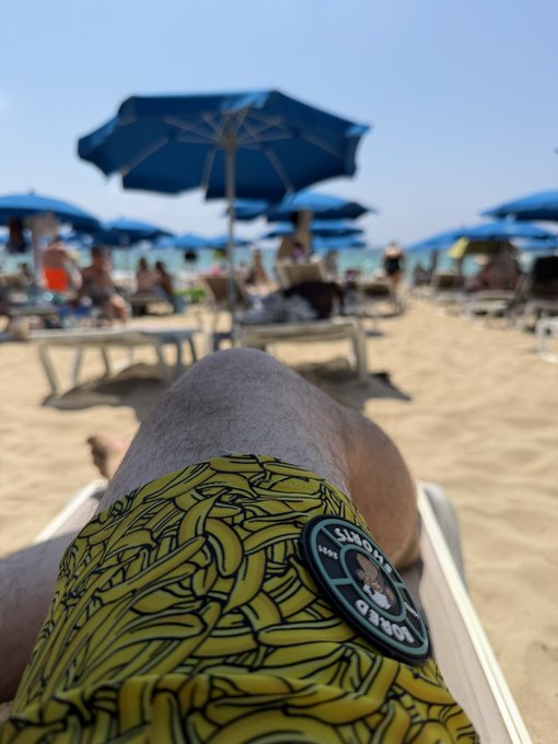 A first-person view from a beach chair shows a person's legs in yellow banana-patterned swim trunks with a 'Lost Tribe Est. 1969' patch, overlooking a sandy beach dotted with blue umbrellas and other beachgoers.