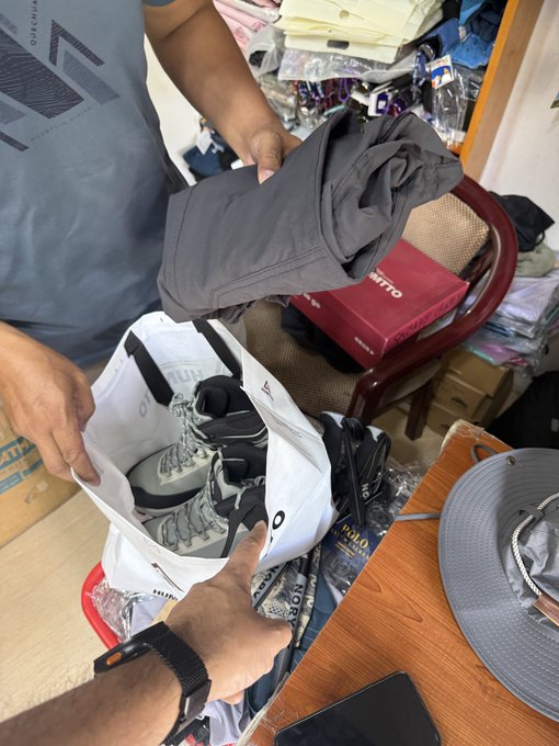 A person holds up dark pants while another hand points into a shopping bag containing shoes, surrounded by other clothing items and a grey bucket hat on a wooden table.