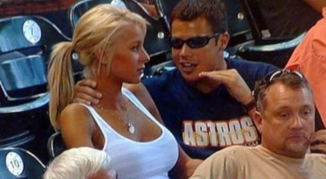 A scene at a sports stadium showing a blonde woman in a white tank top and a man wearing sunglasses and a navy 'ASTROS' t-shirt in conversation.