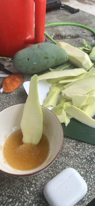 Sliced green mango is dipped into a bowl of orange-brown sauce, with a whole uncut mango visible in the background.