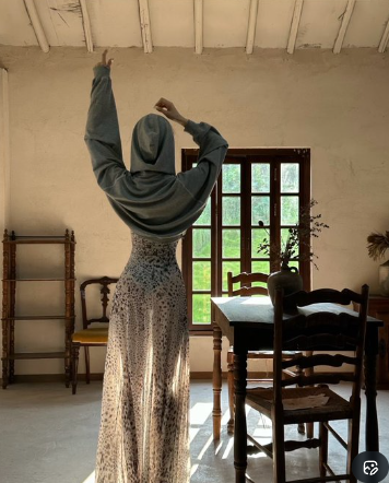 A woman wearing a gray hijab and a long patterned dress stands in a rustic room with her back to the camera, raising one arm in a graceful pose.