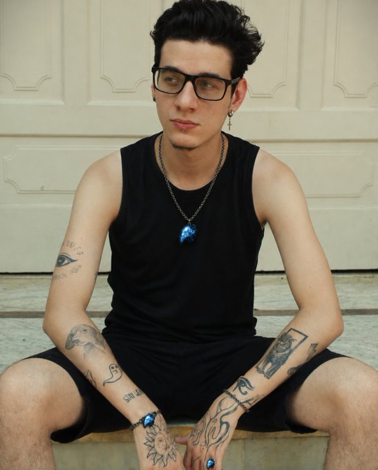 A portrait of a young man with glasses and dark hair wearing a black tank top, featuring various tattoos on his arms and rings on his fingers.