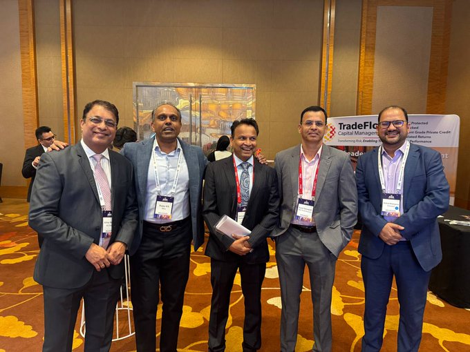 Five men in business suits pose together at what appears to be a TradeFlow Capital Management event, with a banner mentioning "Potential Grade Private Credit Returns and Absolute Performance" visible in the background.