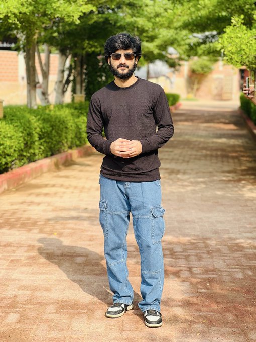 A man with a beard and sunglasses stands outdoors on a brick path, wearing a black sweater, blue cargo jeans, and sneakers, with trees in the background.