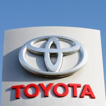 The Toyota logo is displayed on a sign featuring the company's iconic emblem in chrome above the "TOYOTA" name in bold red letters. The image relates to the breaking news about Toyota's announcement of a 1 trillion yen share buyback program.