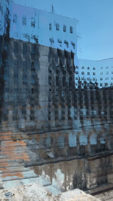 An abstract, distorted image shows the reflection of a building with multiple windows and architectural details against a blue sky, appearing warped and water-like.