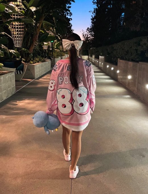 A person walking away from the camera wearing a pink 'POLLY 88' hockey jersey and carrying a plush toy down an illuminated outdoor walkway.