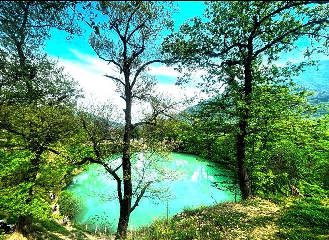 A vibrant turquoise-blue lake or crater surrounded by lush green trees and rolling hills under a bright blue sky.