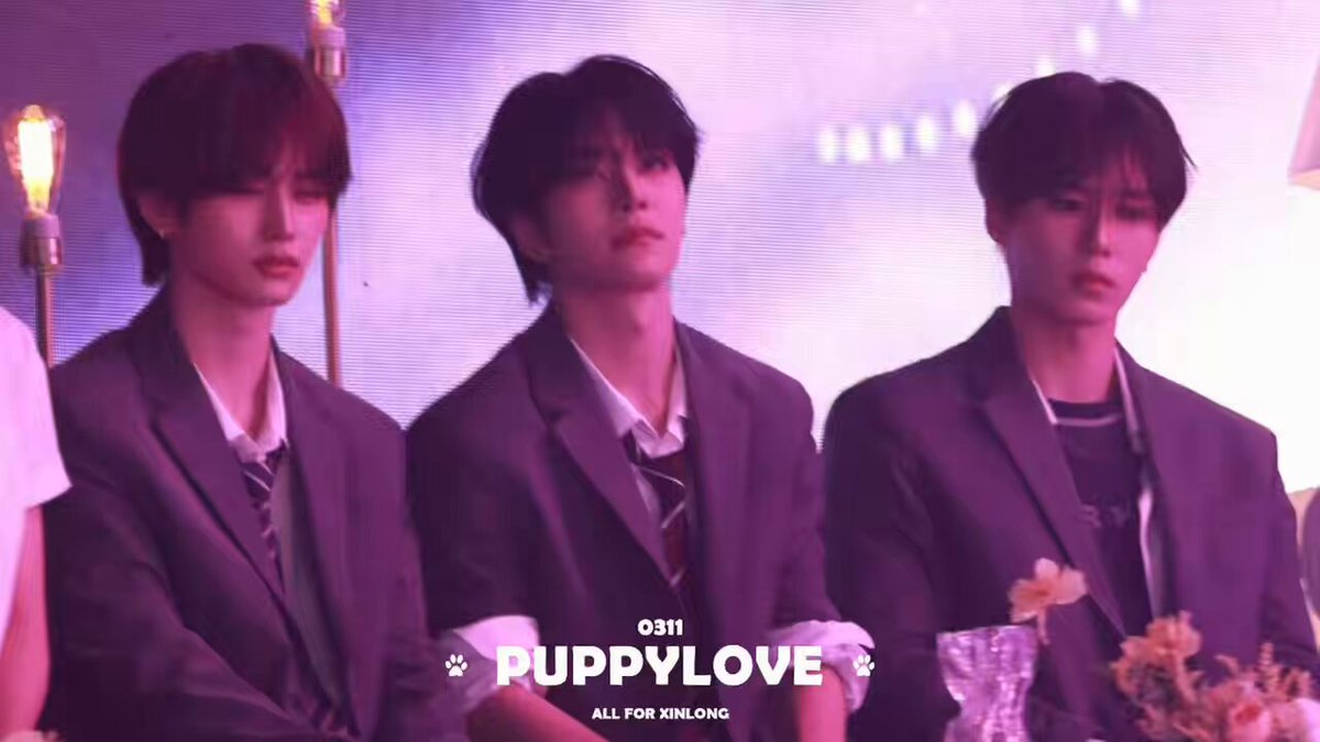 The image features three young men wearing suits, sitting next to each other with their arms crossed. They are all dressed in purple suits and have short hair. The background of the image has a pinkish-purple hue, which complements the color of their suits. There is also a small vase placed on the table between them, adding an elegant touch to the scene.
