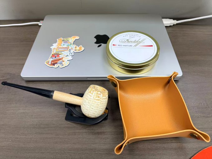 The image features a silver laptop computer with stickers on its surface, including one of a pipe and a cigar tin. The laptop has a sticker depicting a pipe and another showing a cigar tin. The laptop screen displays the text "Scored Brother Shortbird @Wangduanniao's pipe! From now on, I've transformed in a flash—from a rolling buddy to an entry-level pipe buddy." This suggests that the person who owns this laptop has decided to use their pipe as a pipe cleaner or a pipe for smoking.