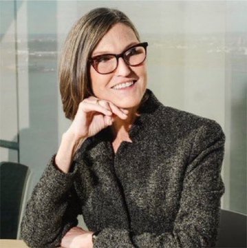 A smiling woman with short brown hair and glasses sits near a window with a waterfront view, wearing a dark textured blazer. The professional portrait doesn't directly depict the post's crypto reference to "$urmom" or SpaceX tokenized stock, but suggests a financial or investment industry context for the commentary.