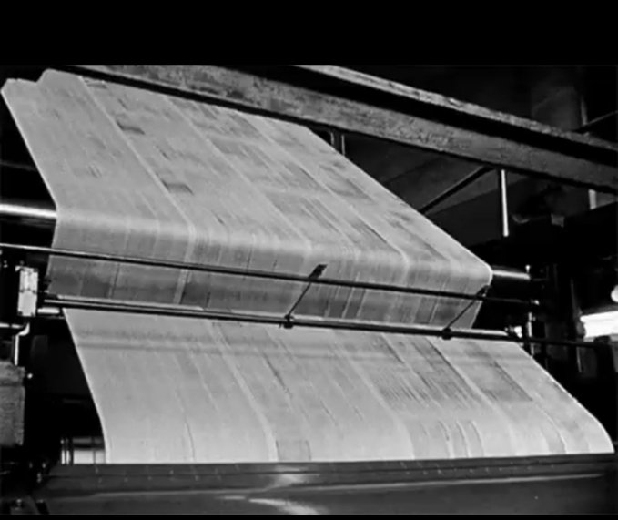 A black and white archival photograph of industrial machinery processing large sheets of paper or fabric through a printing or rolling press.