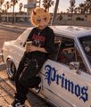 A young boy with blond hair poses next to a white classic car decorated with the word 'Primos' in blue lettering.