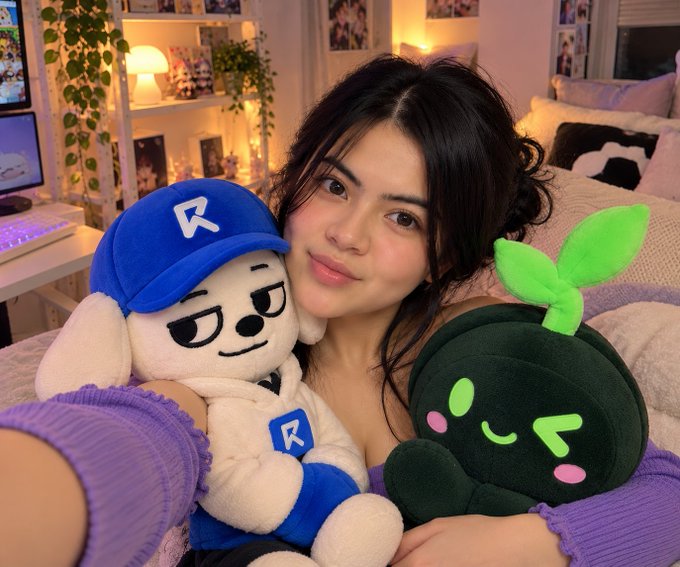 A woman holding two plush toys: a white dog character wearing a blue hat with an 'R', and a dark green plant-like character with a green leaf on its head.