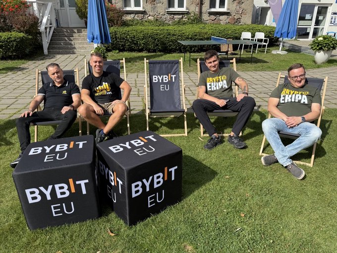 Four men sit in deck chairs and on Bybit-branded cubes outdoors on grass, wearing shirts reading "BYBIT EKRIPA," with one cube and chair displaying the "BYBIT EU" logo.