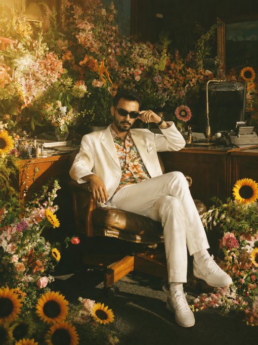 A man in a white suit and floral shirt wearing sunglasses sits relaxed in a leather chair surrounded by an elaborate arrangement of colorful flowers, including sunflowers, in a retro-styled editorial photograph.