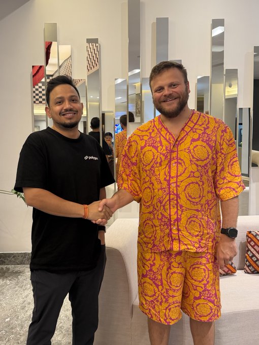 Two men smiling and shaking hands indoors, one wearing a black GoPlus t-shirt and the other in a vibrant yellow and pink patterned shirt and shorts set, with a mirrored wall and white sofa in the background.