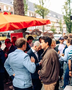 The image shows a group of people standing around an orange umbrella, which appears to be providing shade from the sun or rain. The crowd consists of men dressed in suits and ties, suggesting that they might have attended a formal event or gathering at a café. One man is holding a drink, possibly enjoying a conversation with others nearby. The image also contains text elements such as "@AlpacaHQ" and "@Optimism", which could be related to the event or the people present in the scene.