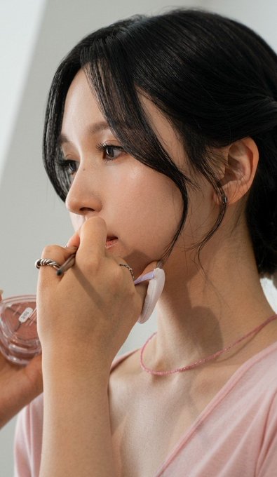 A close-up portrait of a young woman with dark hair pulled back, wearing a pink top, holding her hand near her face with a ring on her finger, holding what appears to be a pink compact.