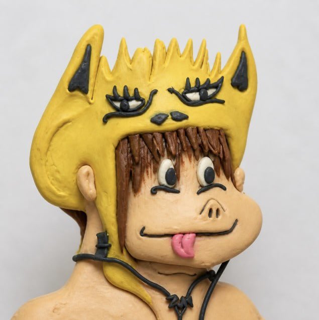 A clay figurine of a character wearing a yellow hood with cat ears, featuring expressive drawn-on eyes and a small pink tongue sticking out.