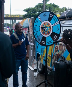The image features a man standing next to a large blue wheel, which appears to be spinning. A truck can also be seen in the background. There are several people around them, including one person holding a pizza box. The scene seems to be set on a street where there is an event taking place or possibly a promotional activity related to BitcoinPizzaDay.