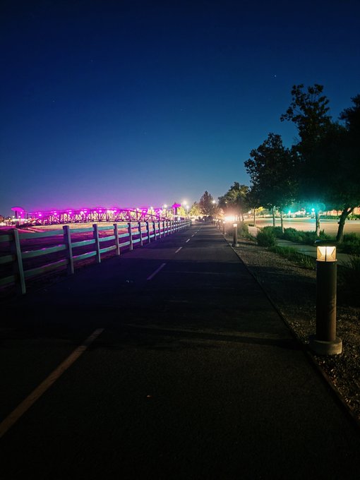 A nighttime photograph of a paved path lined with a wooden fence on the left, leading toward a park illuminated with purple and green lights under a deep blue sky.