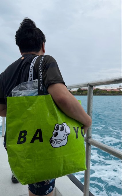 The image shows a man standing near a boat with his arms crossed over his chest while holding a green bag that has "BAY YOU" written on it. The scene appears to be set at a marina or waterfront location, as there is water visible in the background. The man seems to be enjoying his time by the water and possibly taking a break from sailing activities.