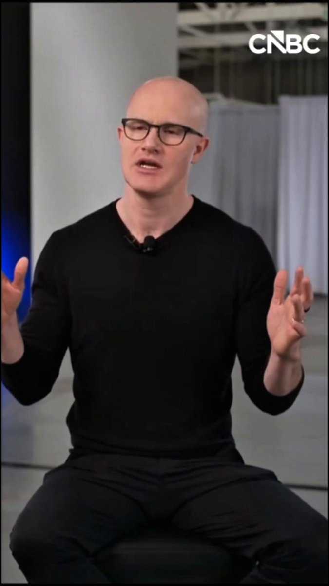 The image features a bald man wearing glasses sitting on a stool or chair with his arms outstretched, possibly giving a speech or presentation. The background of the image shows a white wall with a blue curtain hanging behind him. There are also some text elements in the image that read "base full coinbase" and "cnnbc.com/video/2025/12/...", which may be related to the content being discussed by the man or the context of his speech.