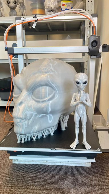 A 3D printer on a shelf next to a silver alien figurine and a large translucent skull with the alien's reflection visible inside, with a printer and filament spool on a shelf above.