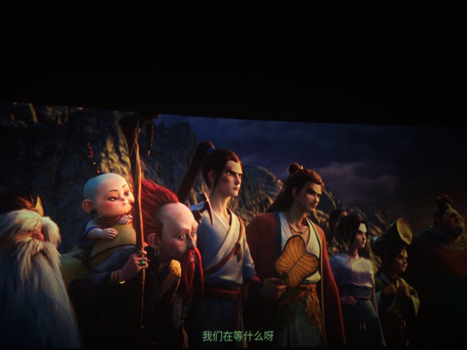 A scene from an animated film shows a group of characters including a monk, a warrior, and various villagers standing together against a dark mountain backdrop, with Chinese subtitles '我们在等什么呀' at the bottom.