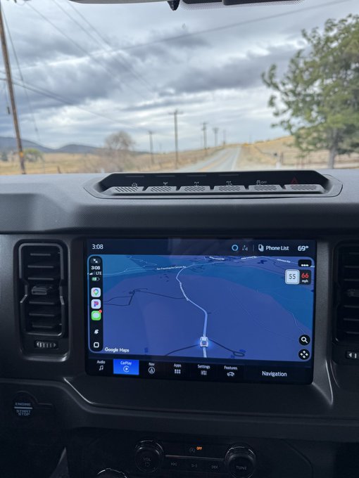 The dashboard of a Ford Bronco shows the infotainment screen running Google Maps navigation, while a rural road and cloudy sky are visible through the windshield.