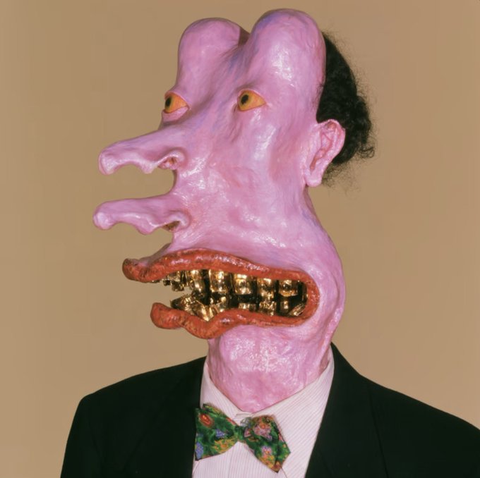 A surreal portrait of a man in a black suit, white shirt, and floral bow tie, wearing a grotesque pink sculptural mask featuring multiple elongated facial features and gold teeth.
