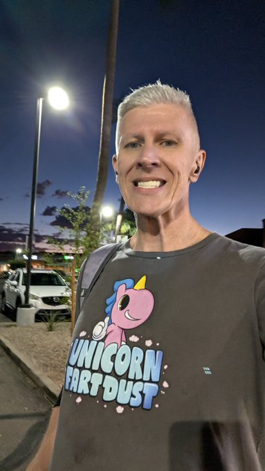 A man with short blonde hair smiles at the camera while wearing a dark gray 'Unicorn Fart Dust' graphic t-shirt at an outdoor nighttime setting.