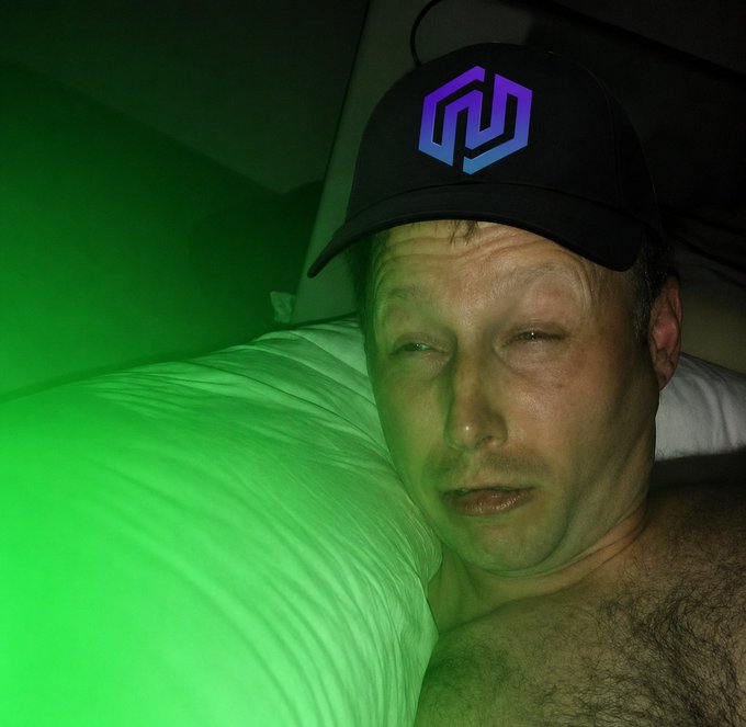 A man wearing a black cap with a purple/blue geometric logo lies in bed bathed in green light, squinting at the camera with a groggy, just-woke-up expression that perfectly matches his "just woke up, what did I miss?" gm message.