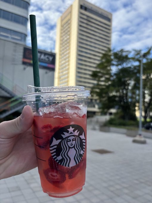 Hand holding a clear Starbucks cup with a green straw containing a pink berry beverage with strawberries, set against an urban backdrop featuring a tall building and a Legacy sign.