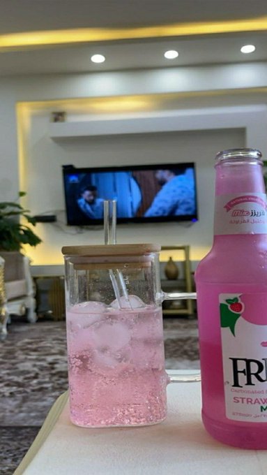 A glass mug filled with a pink iced beverage sits next to a pink bottle labeled 'FR... STRAWBERRY' on a table, with a television playing in the background.