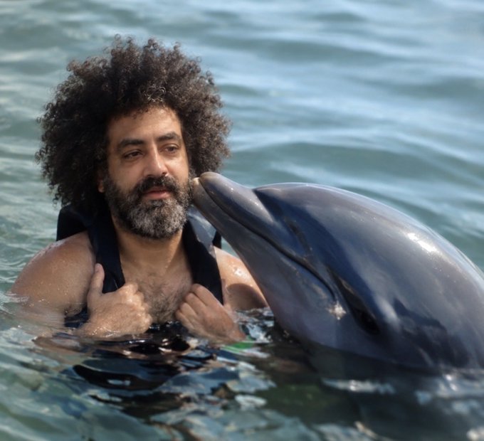 A man with curly hair and a beard poses in the water next to a bottlenose dolphin.