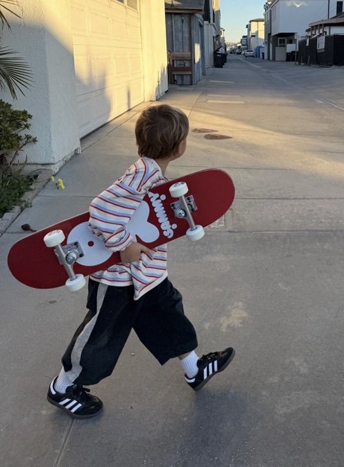 A young boy walking down a residential sidewalk while carrying a red skateboard.