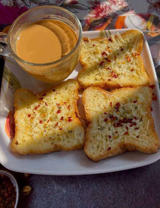 An overhead photograph of a meal featuring three pieces of cheesy garlic toast topped with red pepper flakes, served alongside a glass mug of milk tea on a floral-patterned plate.