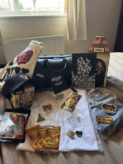 A haul of Doginal Dogs merchandise is spread across a bed, including a t-shirt, cap, mug, sign, and figmas packs labeled "Doginal Dogs" and "Legends Pack," showcasing branded collectibles from the @doginaldogs project.