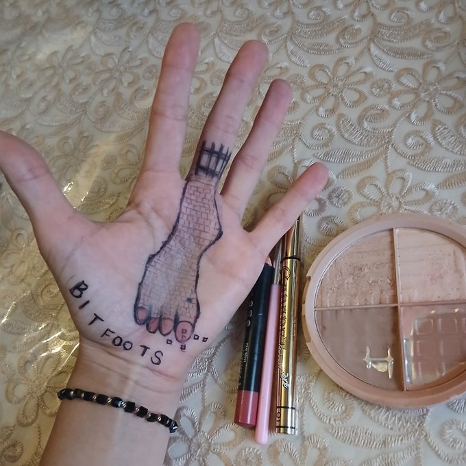 A photograph of a hand with a creative tattoo depicting the Leaning Tower of Pisa across the fingers, accompanied by 'LITTLE BIT FOOTS' text, shown alongside makeup products including eyeliner, lip products, and a contour palette.