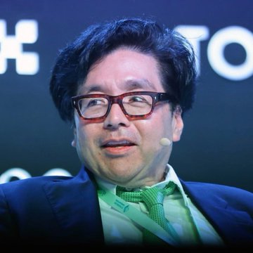 A bespectacled man wearing a dark suit and green patterned tie speaks into a headset microphone, likely at a financial or crypto conference. The image likely depicts Tom Lee, the leader of Bitmine mentioned in the accompanying post about the company's significant ETH purchase from Kraken.