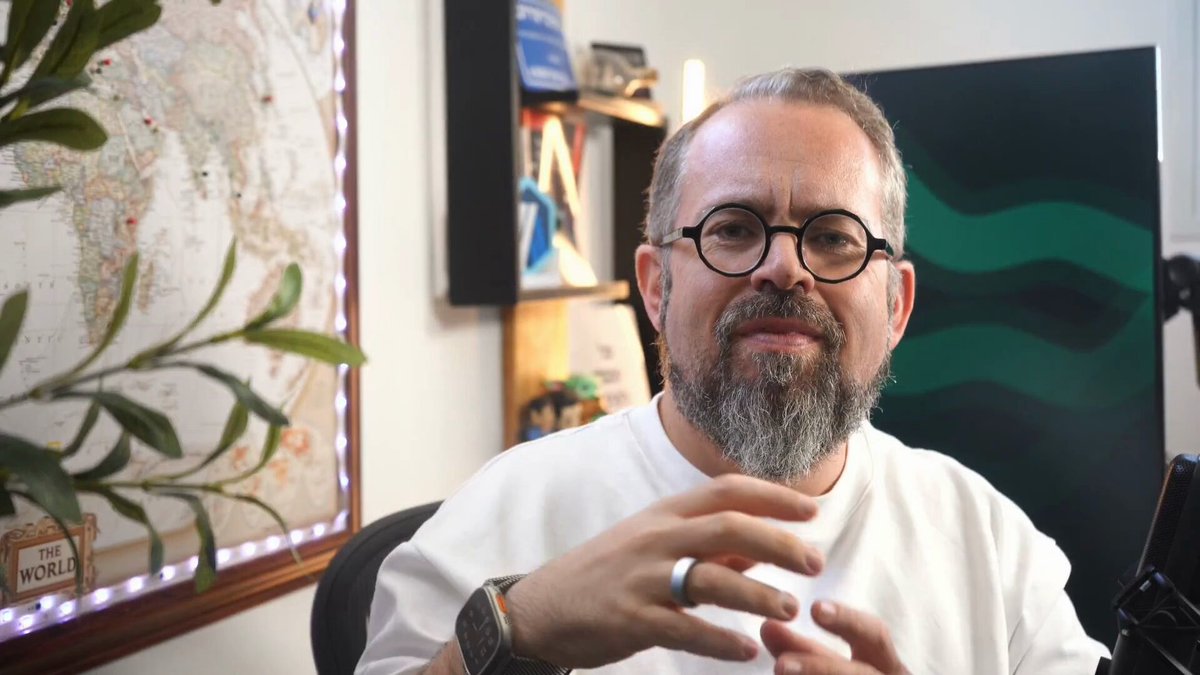 The image shows a man wearing glasses, sitting at a desk with his hands together as if he's about to speak or make a gesture. The background features a map of South America along with some books scattered around him. There are also two potted plants in the room - one on the left side and another on the right side. The image does not contain any text, but it seems to be related to SQL databases as there is a post about SQL databases that includes a code for an SQL database called @arbitrum.