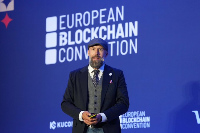 A bearded man in a flat cap, vest, and tie speaks on stage at the European Blockchain Convention, with "KUCOIN" and "EUROPEAN BLOCKCHAIN CONVENTION" visible in the backdrop, illustrating the post's feature of @F_Gregaard presenting Cardano's role in solving institutional trust in digital finance.