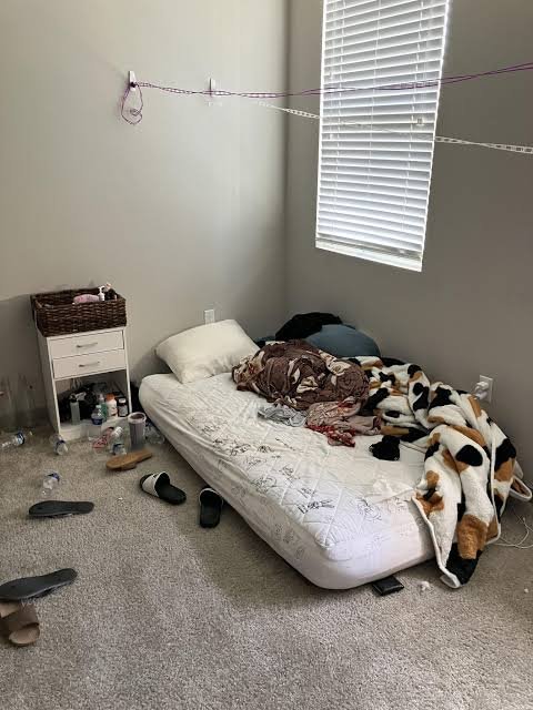 A messy bedroom shows a mattress on the floor covered with rumpled blankets, surrounded by scattered belongings, bottles, and shoes on the carpet.